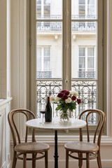 Fototapeta premium Parisian kitchen nook with round marble bistro table, cane chairs, wine bottle, and bouquet by tall window overlooking wrought-iron balcony