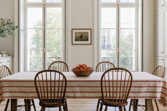 Parisian kitchen dining corner featuring a long farmhouse table with red and cream striped linen cloth and Windsor chairs, bright light from tall windows