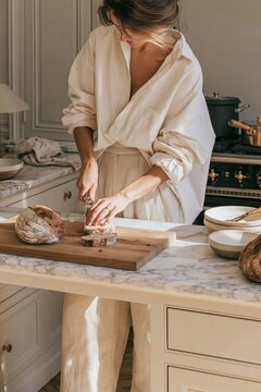 Young woman in oversized cream shirt and wide-leg linen trousers slicing bread on wooden board in elegant Parisian kitchen with marble countertop