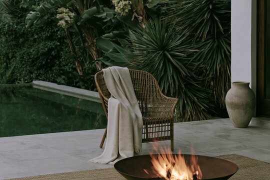 Wool throw draped over rattan armchair beside a glowing fire pit on a Tropical Modernism terrace surrounded by lush greenery at dusk