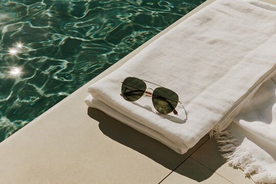 Sunglasses resting on a folded white linen towel beside a turquoise pool in a Tropical Modernism villa under warm midday light