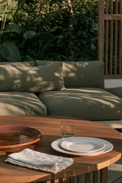 Stack of folded linen napkins beside white plates and a wooden bowl on a low round teak table in a tropical modernism terrace setting