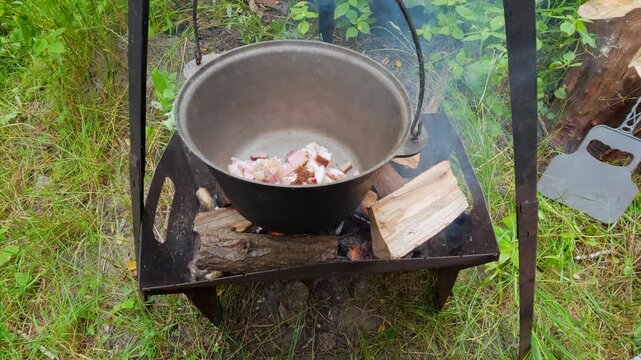 Man adding chopped bacon into cauldron over campfire for outdoor picnic cooking lifestyle