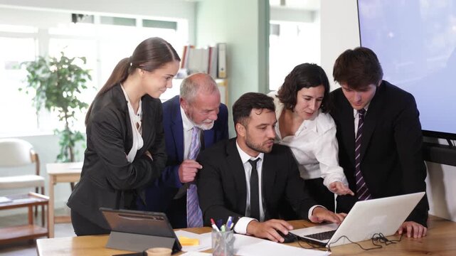 Group of business people discuss business strategies and planning work together, everyone looking at laptop by man and laughing in bright corporate office