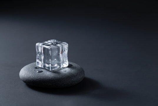 A clear ice cube rests on a smooth, round black stone against a dark background, creating a minimalist and serene composition.
