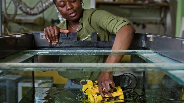 Young adult African American female scientist lifting yellow underwater drone from testing tank, camera tilting up. Footage is perfect for marine engineering and robotics technology marketing