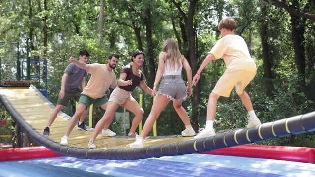 Men women and children attempt to cross unstable swinging bridge suspended over large trampoline