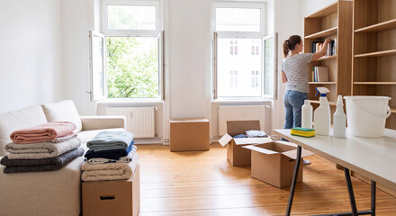 Naklejka premium Young woman organizes wooden shelving units near stacks of moving cardboard boxes and folded blankets inside a sunlit empty living room apartment