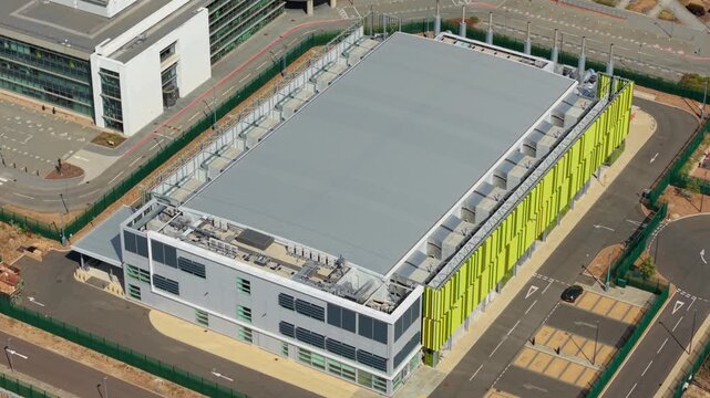 Aerial view of a modern hyperscale AI data centre campus featuring rows of indirect evaporative cooling units on the roof and green architectural facade panels in Harlow, England, United Kingdom.