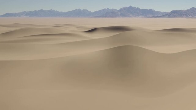 Dry sand dunes cover the ground in a vast desert. The scenery shows rolling shapes and lines in the sand. Mountains rise in the background under a clear sky.