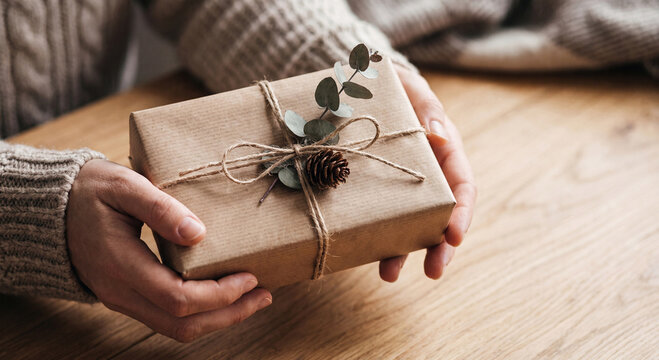 Hands holding beautifully packaged gift box in brown craft paper with rustic string and green eucalyptus leaf on wooden background