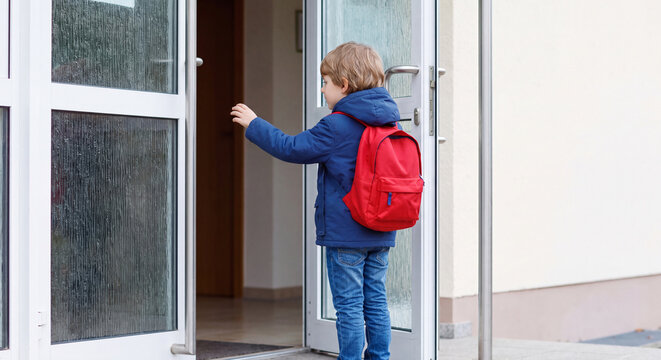 A cute young schoolboy wearing a blue jacket and red backpack pushes open the glass front doors to enter a modern school building in the morning