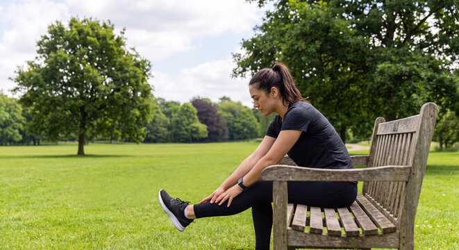 Sporty young woman in black activewear performing hamstring stretch on a wooden bench in a scenic green park during daytime workout session