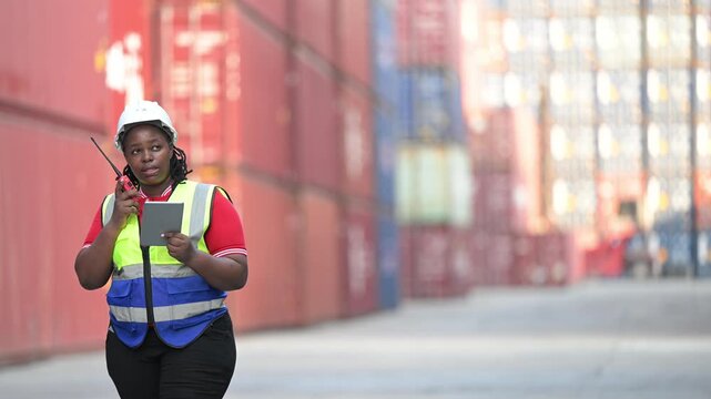 One female logistics worker uses a two way radio and hand signals to direct cargo operations at a shipping container yard. Concept of logistics, freight transport, communication, and supply chain.