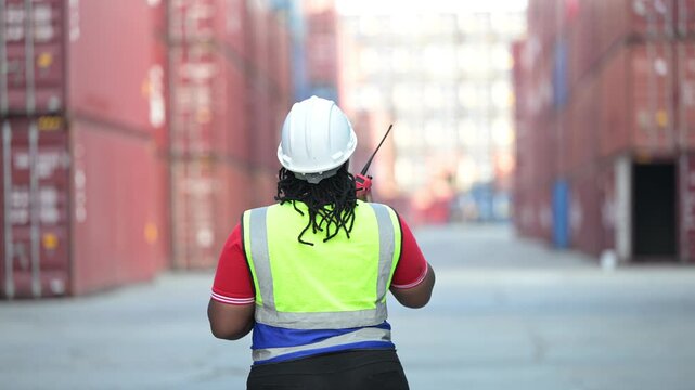 One female logistics worker uses a two way radio and hand signals to direct cargo operations at a shipping container yard. Concept of logistics, freight transport, communication, and supply chain.