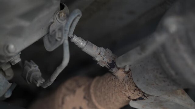 An oxygen sensor with electrical wiring screwed into a rusty car exhaust pipe underneath the chassis.