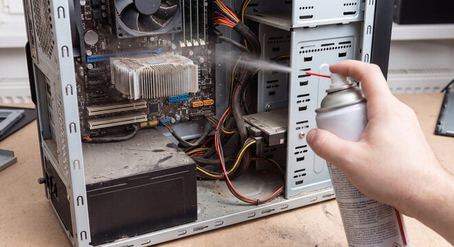 Hand spraying pressurized air from aerosol can onto dusty heatsink and motherboard inside open computer case during preventive cleaning