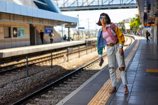 A smiling woman waits for a train and uses a smartphone at a train station. A young woman holds a smartphone and stands at a train station.