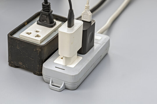 Close-up of an old, dusty electrical power strip and extension outlet overloaded with multiple phone chargers, adapters, and tangled cables all plugged in simultaneously