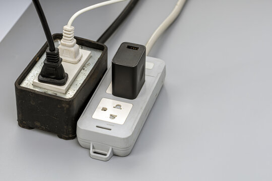 Close-up image of two worn and dusty electrical power strips overloaded with multiple plugs, a black USB adapter, and tangled cables, highlighting the danger of electrical overload and fire hazard