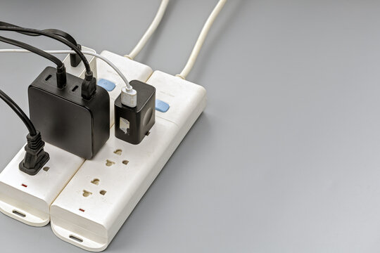 Close-up view of a white power strip crowded with multiple black cables, USB wall chargers and power adapters plugged in simultaneously, illustrating electrical overload and fire hazard concept