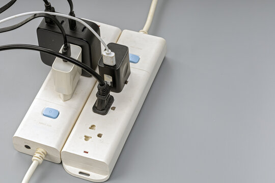 Close-up of a white electrical power strip overloaded with several black plug adapters, chargers, and tangled wires, illustrating electrical hazard and overload risk on a gray background