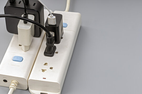 Close-up view of a white electrical extension power strip overloaded with several black and white phone chargers, adapters, and cables plugged into every available outlet