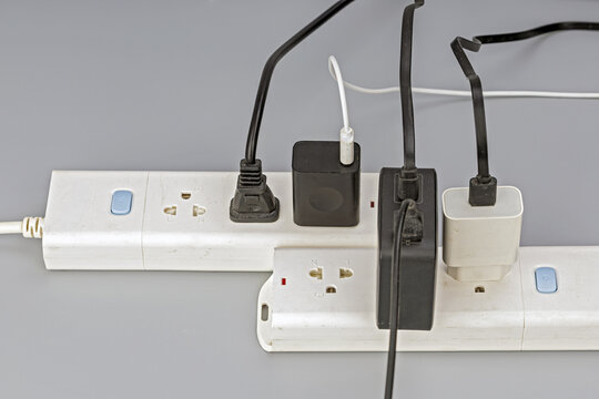 Close-up view of two white electrical power strips crowded with multiple black and white power cables, USB chargers, and adapter plugs, illustrating electrical overload and cluttered wiring against