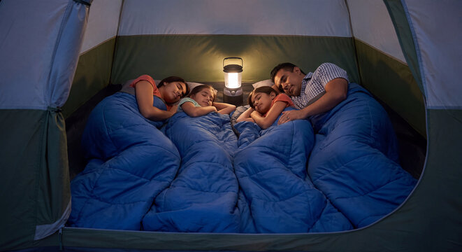 Family with young children resting asleep in warm blue sleeping bags inside a camping dome tent illuminated by lantern light at night