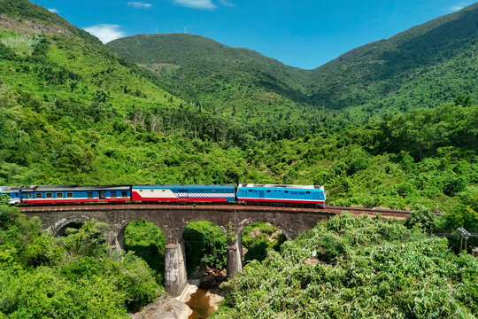 Blue passenger train crossing a high stone arched bridge surrounded by vibrant green tropical mountains under a clear sky during a bright daytime journey