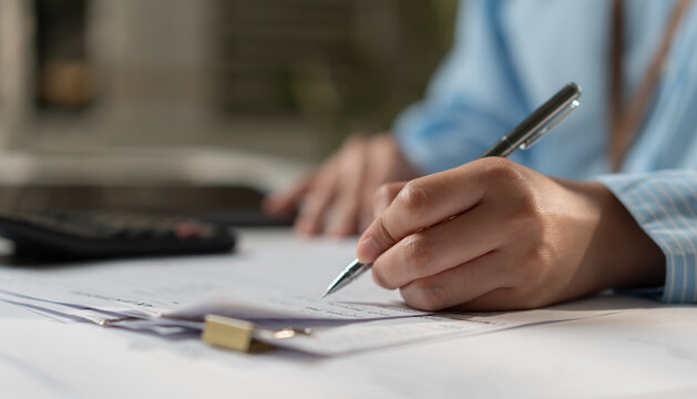 Businesswoman writing on financial documents with a pen and calculator at office desk, representing accounting, budget planning, paperwork, expense tracking and financial management.