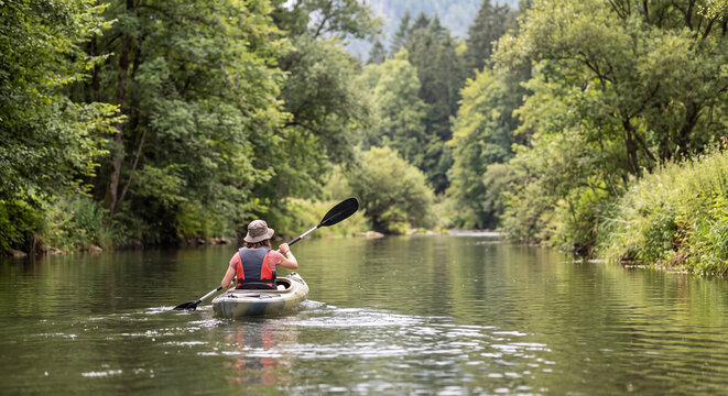 Rear view of an active person kayaking along a serene calm river surrounded by dense lush green trees in summer nature