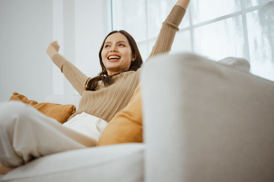 A woman stretches both arms while relaxing on a sofa at home, embracing relaxation, comfort, mindfulness and wellness in a peaceful morning lifestyle where calm breathing deepens her relaxation.