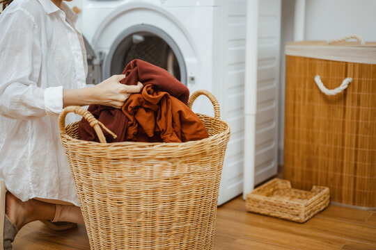 A woman sorts clothes into a laundry basket beside the washing machine, organizing fabric, garment and textile care for a clean home routine where each laundry load improves housekeeping efficiency.