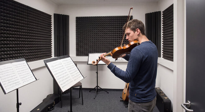 A male violinist practices playing classical violin facing empty music stands inside a modern soundproof music recording and rehearsal studio