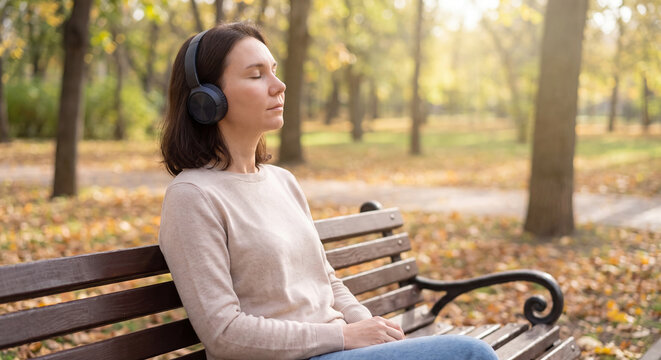 Young woman sitting on a wooden bench in an autumn park with closed eyes listening to peaceful music through modern headphones enjoying fall nature