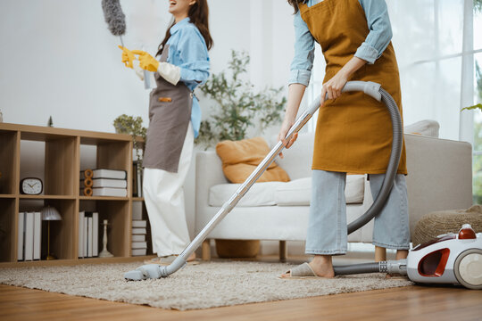 Two women use a vacuum cleaner across a rug, combining cleaning, housekeeping and teamwork to remove dust, refresh the home, protect the sofa and complete household chores with careful vacuum care.