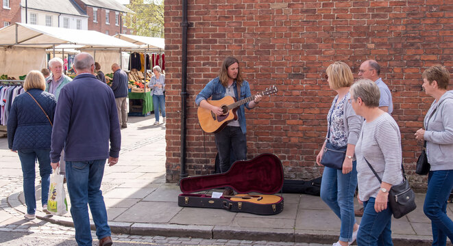 Young male busker sitting on a stool playing acoustic guitar with an open guitar case for coins as pedestrians walk past brick buildings