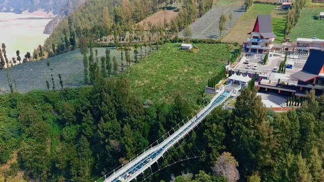 Aerial view of Jembatan Kaca glass bridge surrounded by lush green mountains and modern architecture in Kecamatan Sukapura, Jawa Timur, Indonesia.