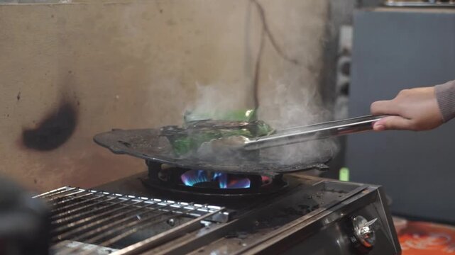 Close-up of street food cooking on a hot griddle over gas flame, with steam, metal tongs, and fresh food preparation in a busy kitchen.