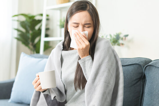 Sick, hurt or pain asian young woman have a fever, flu sore throat, drinking a mug of warm water, use tissues paper sneezing nose, runny while sitting rest on sofa at home. Health care on virus person