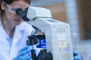 A female biotechnology researcher wearing a white laboratory coat, hairnet and blue nitrile gloves reaching up toward the objective turret inside a busy medical diagnostics laboratory. © ultramansk