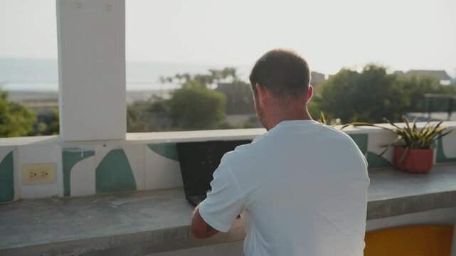 Man typing on a laptop at a seaside terrace during a beautiful sunset, living a digital nomad lifestyle
