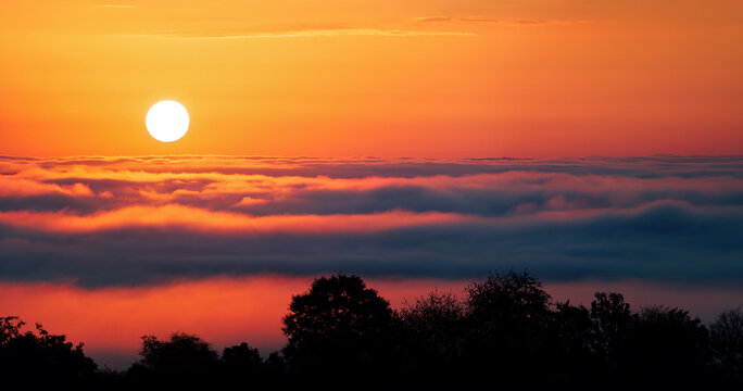 Calm sunrise and deep orange sky over wavy fog or clouds. Fascinating weather phenomenon with beautiful vibrant colors.