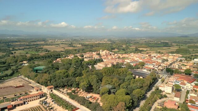 Aerial view of the historic town of Peralada surrounded by lush green trees and agricultural fields under a clear blue sky in Peralada, Catalonia, Spain.