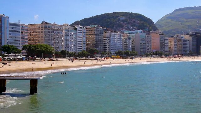 A scenic panning view of Copacabana beach in Rio de Janeiro on a sunny day, featuring sandy shores, blue ocean waves, beachfront buildings, and majestic mountains in the background.