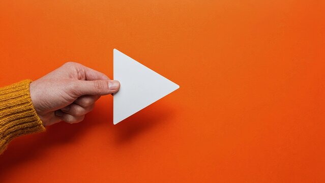 Fingers holding a white play button shape on a textured orange surface creating an engaging visual direction for media content.