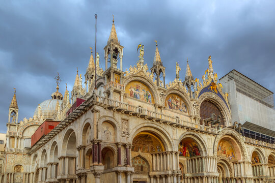 View of St. Mark's Basilica with its ornate facade, golden mosaics, and intricate marble arches under a dramatic cloudy sky in Venice, Veneto, Italy.