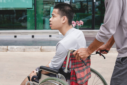 Young Asian man in a wheelchair pushed by a caregiver outdoors in front of a modern building, concept of accessibility, mobility, and inclusive lifestyle.
