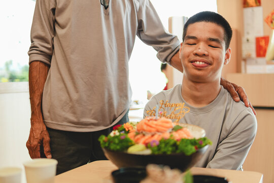 Happy young Asian man in a wheelchair closing eyes and making a wish for his birthday with father, salmon sashimi cake on table, family love, inclusive life, and celebration.